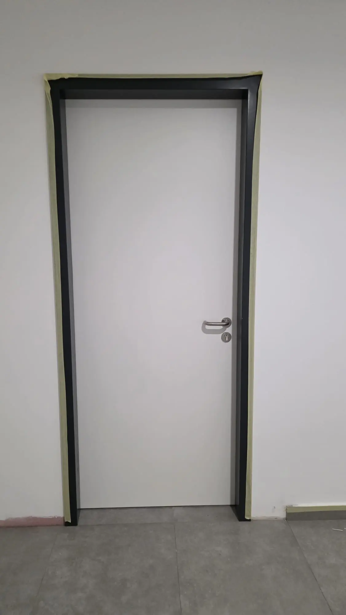 Bijela vrata s crnim okvirom i srebrnom kvakom tijekom renovacije, s maskirnom trakom oko okvira.