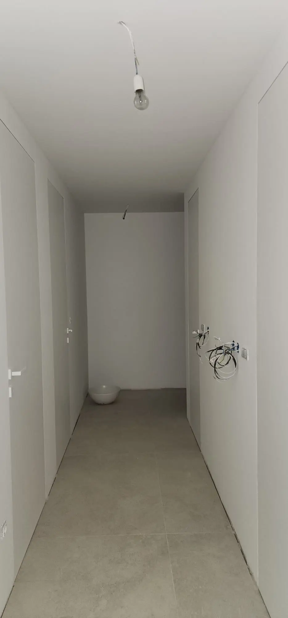 Hodnik u izgradnji s bijelim zidovima, sivim podnim pločicama i izloženim električnim instalacijama