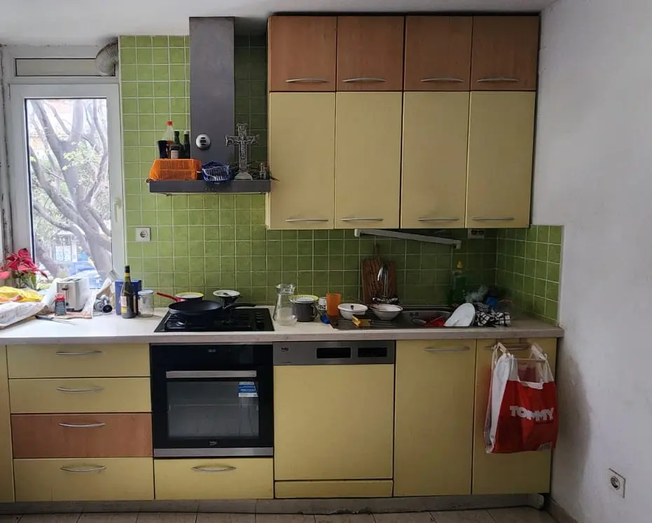 Stara kuhinja sa zelenim zidnim pločicama, žutim ormarićima i prozorom s pogledom na drveće.