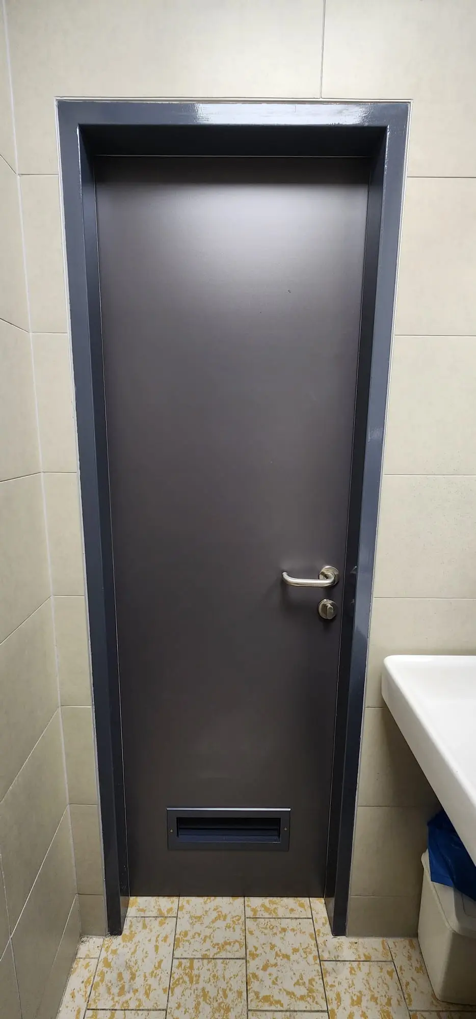 Tamna siva vrata s metalnom kvakom i otvorom za poštu, okružena bež pločicama i žutim podom.