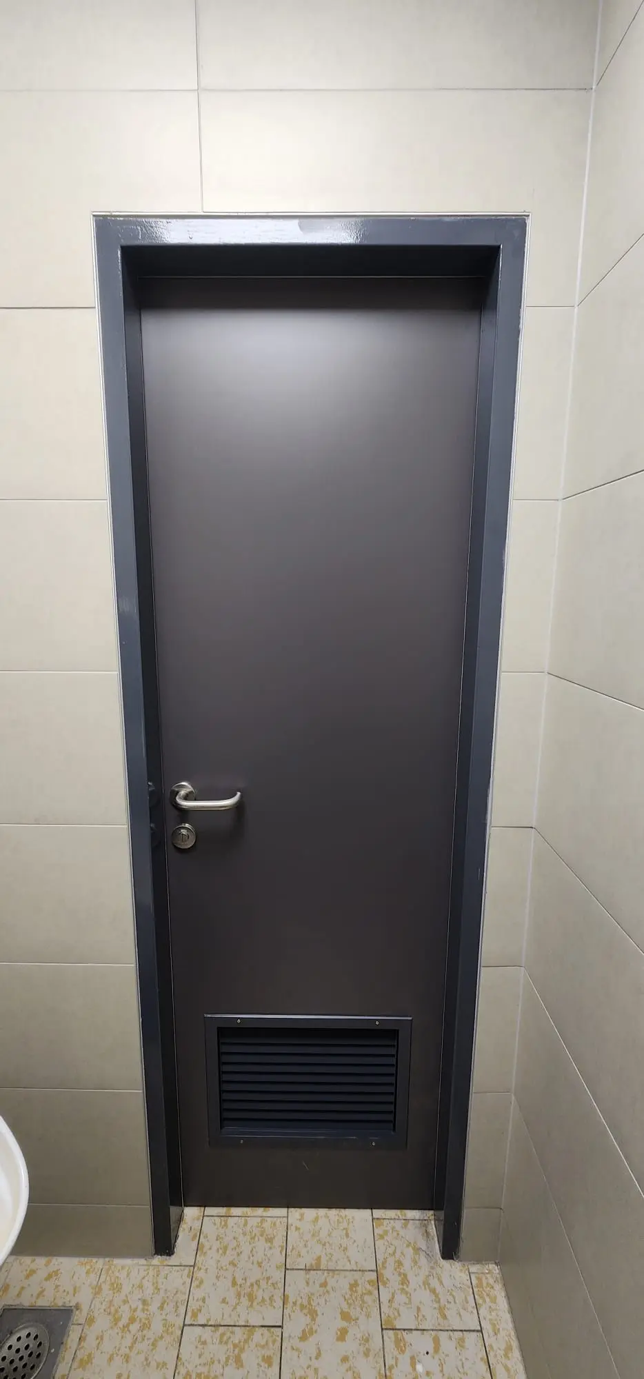 Tamna siva vrata s metalnom kvakom i ventilacijskom rešetkom u prostoru s pločicama.